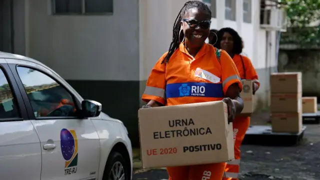 Mulher vestida com uniforme da prefeitura do Rio de Janeiro, carrega uma caixa contendo uma urna eletrônica