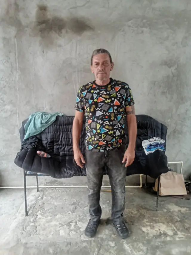 Lázaro Romero León mira a cámara de pie frente a una silla alargada, con jeans gris oscuros y una camiseta estampada, en un patio de cemento en Tapachula, Chiapas, México.