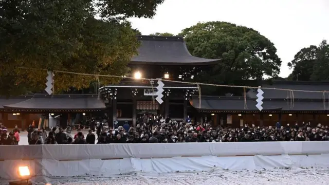緑に囲まれた神社の門に「南神門」と書かれ、白いビニールシートが敷かれた賽銭(さいせん)入れの前に大勢が集まっている。上にしめ縄がかけられている。