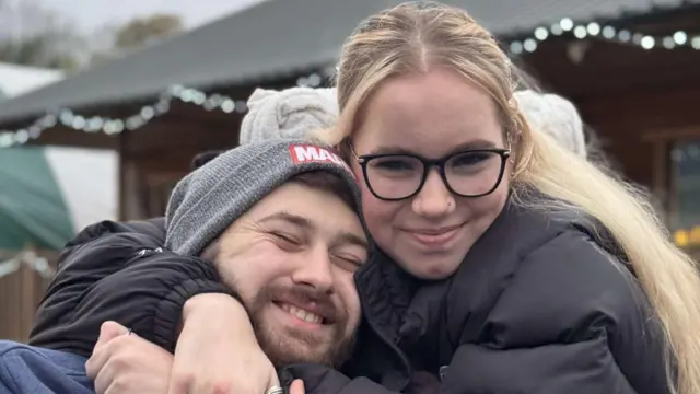 Megan Dixon abraçando o namorado. Megan é loira e está usando óculos com armação preta e um casaco preto. Oliver usa uma touca cinza e barba e está de olhos fechados sorrindo. 
