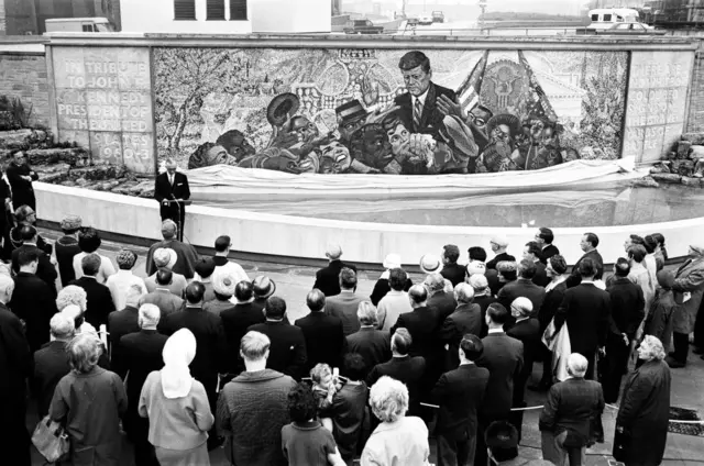 Multitudes reunidas alrededor de un nuevo mosaico conmemorativo en memoria de John F. Kennedy. Es una imagen en blanco y negro del homenaje en los Jardines Kennedy de Birmingham.