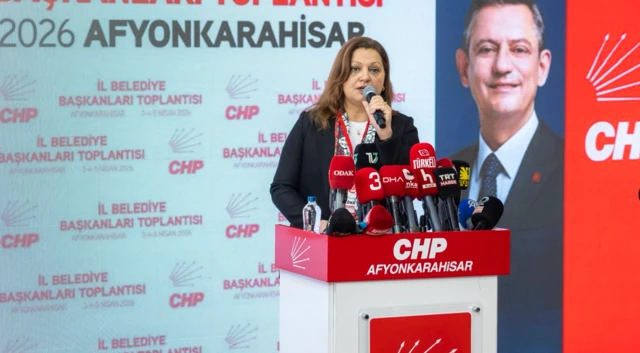 Çok mikrofonlu bir kürsüde bir kadın belediye başkanı konuşuyor. Arkada Özgür Özel'in fotoğrafı var