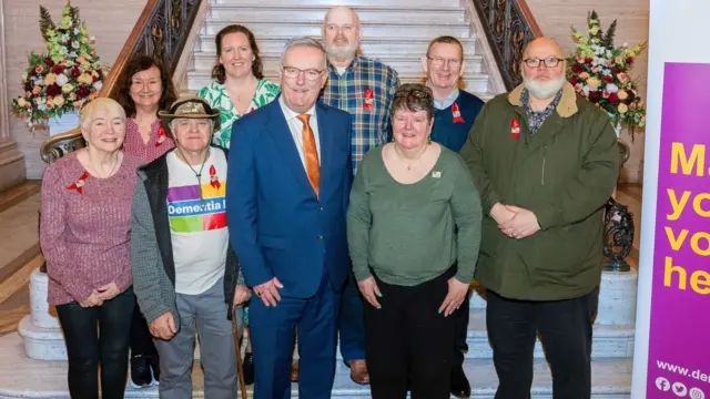 Oito membros do Dementia NI ao lado de Mike Nesbitt ao pé de uma grande escadaria 