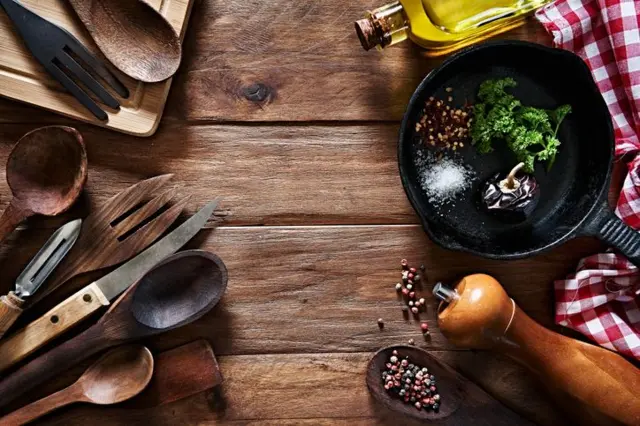 Ahşap kaşık ve çatallar solda sağda biberlik küçük tavada yeşillikler ve zeytinyağı görünüyor. Hepsi rustik bir masanın üzerinde.