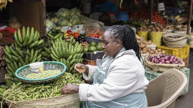 Une femme, vendeuse, sur un étal de marché au Kenya avec des pois verts, des bananes vertes et d'autres produits d'épicerie.