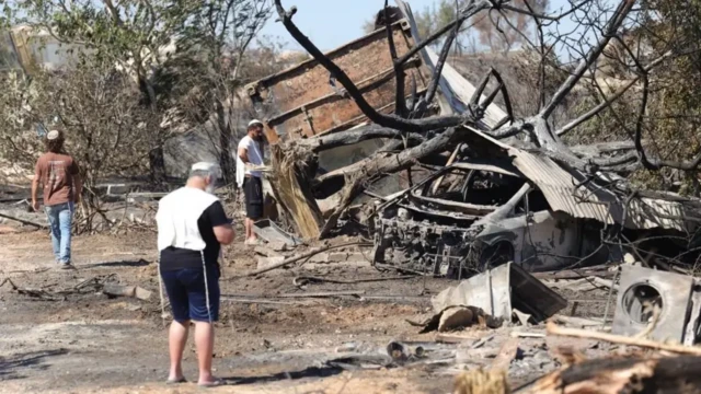 یک خودروی سوخته و بقایای یک خانه و درختان و زمین خشک، سه یهودی در حال بازدید