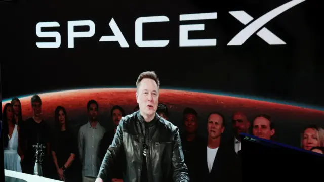 Elon Musk aparece por meio de transmissão de vídeo, vestindo uma jaqueta de couro preta e discursando em um pódio, durante a estreia de sua empresa SpaceX na bolsa de valores Nasdaq, em junho.