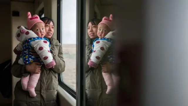 Una china y carga a su bebé en brazos junto a la ventana de un tren. 