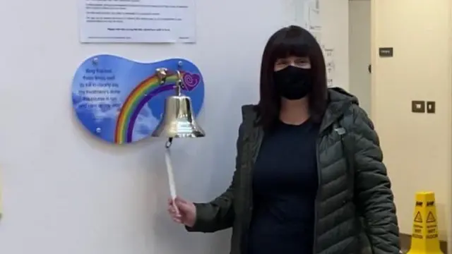 Trish ringing a silver bell which signifies the end of her cancer treatment. She has short dark hair and is wearing a green coat, black top and black mask.