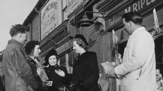 Тэтчер, в черном пальто и шляпке, разговаривает с избирателями на улице. Справа от нее две жунщины и один мужчина, слева мужчина в белой куртке.