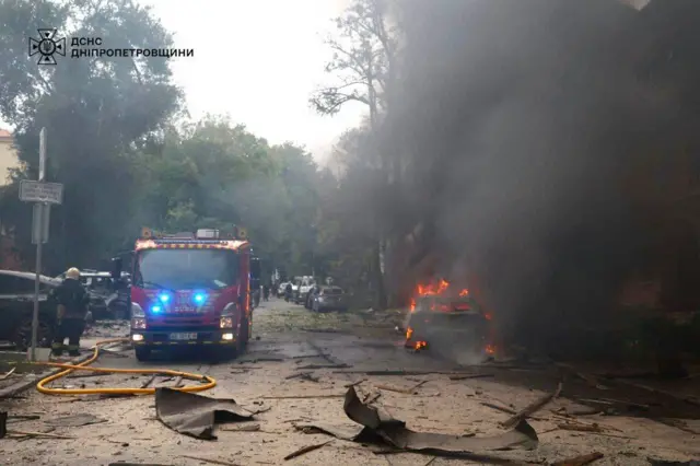 Вулиця після обстрілу, уламки та горить машина. Стоїть машина рятувальників