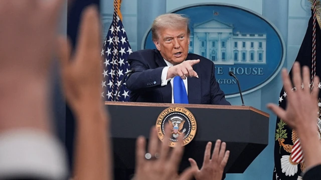 Trump bir kürsüde, elini kaldırmış gazetecilere söz veriyor