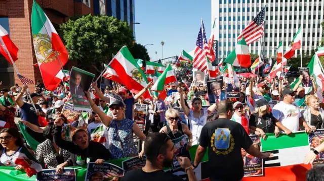 晴天の街の一角に、イラン革命前の旗や、さまざまなプラカードを持った人たちが集まっている