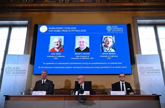 Três membros do comitê do Prêmio Nobel sentam-se a uma mesa de madeira, atrás de microfones, em uma coletiva de imprensa. Os três homens são Olle Eriksson, presidente do Comitê Nobel de Física, Hans Ellegren, secretário-geral da Real Academia Sueca de Ciências, e Goeran Johansson, membro do Comitê Nobel de Física. Atrás deles, há uma tela com fundo azul e fotografias dos três vencedores do prêmio deste ano.