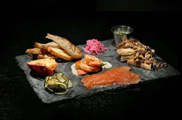 Une planche de produits fumés et salés, comprenant des moules en escabèche, des crevettes épicées, de la queue de homard fumée et du saumon. 