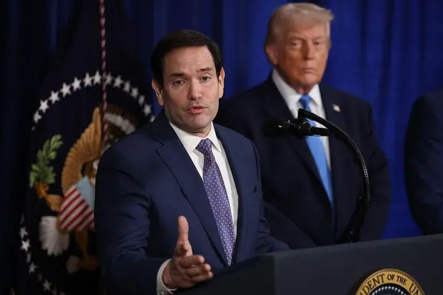 Sekretarz stanu USA Marco Rubio odpowiada na pytania podczas konferencji prasowej przy mównicy, na tle której widnieją insygnia prezydenckie i prezydent Donald Trump. Obaj ubrani są w ciemnoniebieskie garnitury i niebieskie wzorzyste krawaty.