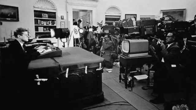 A black and white photo show US President John F Kennedy address di nation from di White House during di Cuban Missile Crisis on October 22, 1962