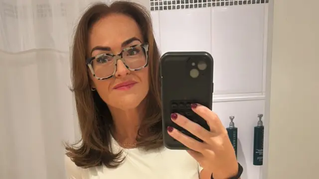 Una mujer con pelo lacio castaño, una camiseta blanca, anteojos y lápiz labial rosa se toma un "selfie" con un teléfono ante el espejo