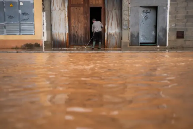 Una persona barre el lodo de la puerta de su casa en una calle absolutamente anegada de agua.