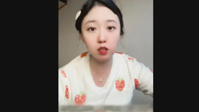 一位名叫李宜雪的年轻中国女性面对镜头，身穿印有草莓图案的白色上衣，涂着红色唇膏，头发紮起并用发夹固定。