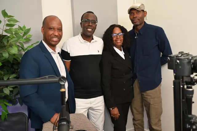 Umunyamakuru wa BBC ari kumwe n'abatumirwa be bo mu kiganiro, bifotoje bahagaze inyuma hari urukuta, ku ruhande hari ururabo rw'icyatsi kibisi.