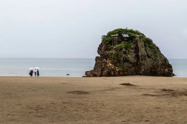 Tres mujeres pasean por la playa en la que se puede ver una gran formación rocosa con un templo