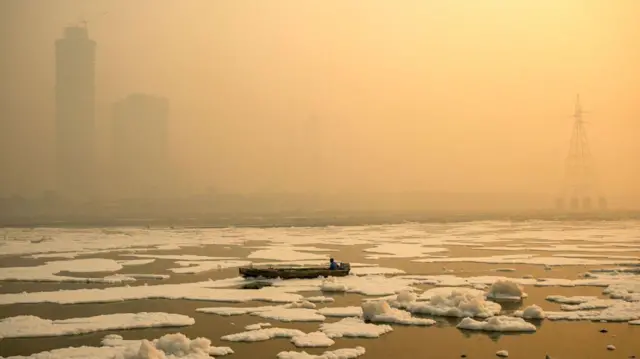 Um homem no rio coberto de espuma tóxica, com skyline de Delhi ao fundo