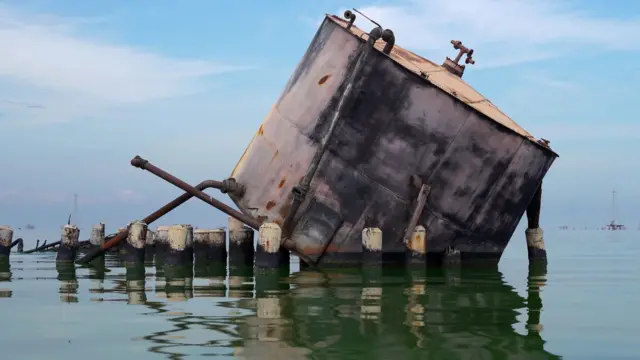 Un gran tanque metálico redondo en desuso, con tuberías que sobresalen, se encuentra a nivel del agua. Está inclinado, de modo que parte de la base y del lateral derecho están sumergidos. Delante del tanque sobresalen del agua postes cortos y gruesos, con la parte superior de color amarillento.