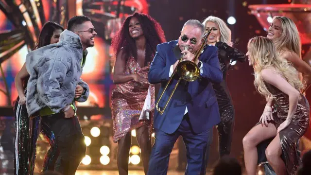 Willie Colón tocando el trombón y rodeado de bailarines
