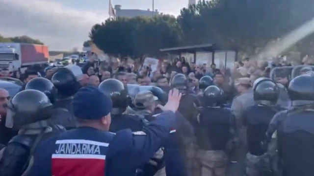 Jandarma yazılı üniformalı bir personel arkası dönük, önünde onlarca çevik kuvvet polisi var, önlerinde siviller flu şekilde görünüyor. 