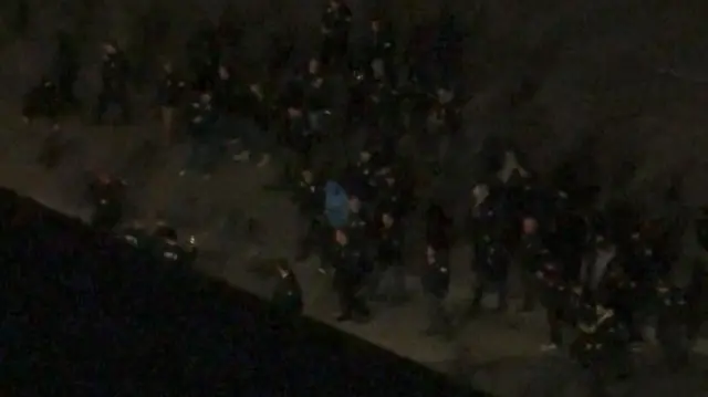 暗い屋外写真。黒い服装の人物が大勢いる中央に、灰色の服装の人物が見える