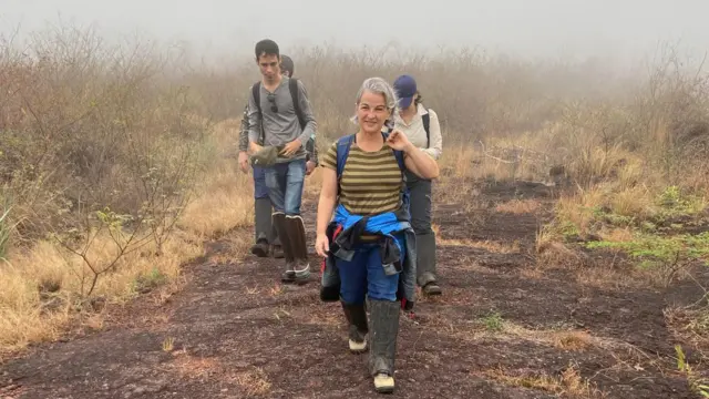 Ana sorrindo ao caminhar em área com arbustos; há outras pessoas caminhando atrás dela