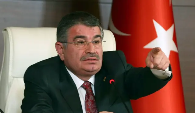 İdris Naim Şahin üzerinde bir takım elbiseyle konuşurken elini kaldırmış, birini işaret ediyor. Arkasında Türk bayrağı ve önünde bir mikrofon var