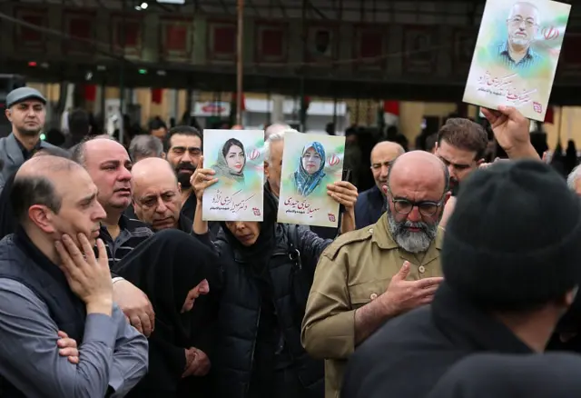 Civiles en una calle iraní, llorando por sus seres queridos. Una mujer sostiene fotos de dos mujeres.