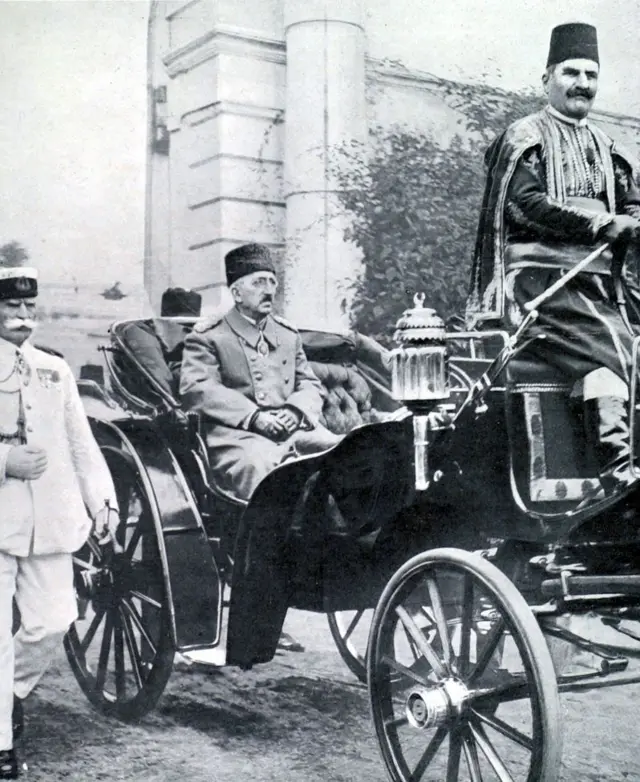Mehmed VI, lleva un abrigo, está sentado en carruaje que transita por la calle. A su lado, camina un funcionario vestido de blanco