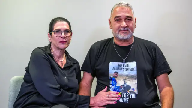 イスラエル人人質ラン・グヴィリ氏の写真がプリントされた黒い半袖Tシャツを着た父親の隣に、グヴィリ氏の母親が座っている。グヴィリ氏の母親はTシャツの息子の写真に右手を添えている。両親は口を閉じ、カメラを見ている