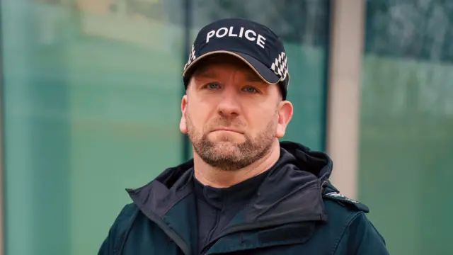 Un hombre con una gorra de béisbol negra de policía mira directamente a la cámara. Lleva una camiseta y una chaqueta negras. Tiene una barba castaña y pulcra.