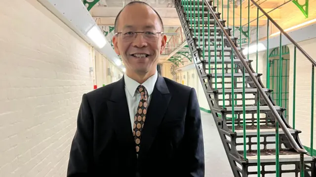 Un hombre chino con traje negro, camisa blanca y corbata estampada amarilla y negra sonríe a la cámara. Detrás de él hay una escalera de hierro verde oscuro y paredes de ladrillo pintadas de color crema.