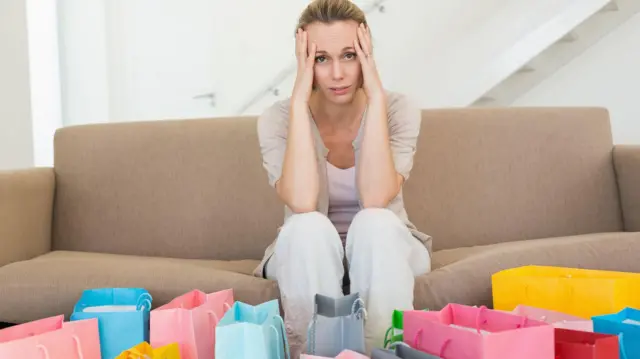 سيدة تجلس على أريكة وأمامها عدد كبير من حقائب التسوق، وتبدو على وجهها علامات الندم