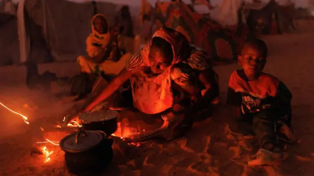 A woman in a refugee camp cooks at an open fire with the light from the flames illuminating her face. A child sits to her left.