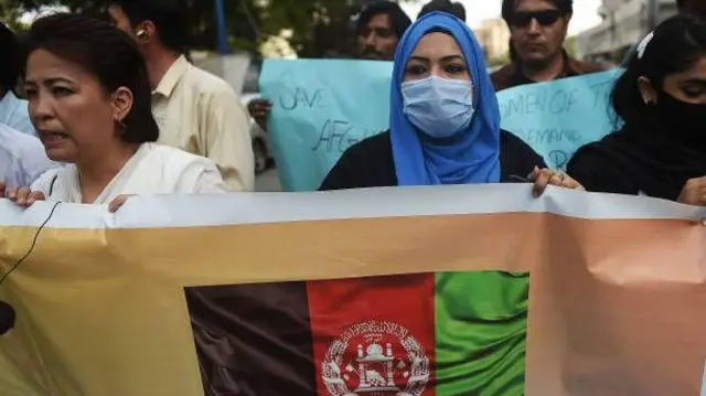 نمایی از سه زن که پلاکاردی با پرچم افغانستان را در دست دارند. در میان یک زن افغان دیده می‌شود که چادر آبی دارد و صورتش را با ماسک آبی پوشانده است. در عقب شان نیز چند مرد دیده می شود که آن‌ها نیز شعارهایی در دست دارند.