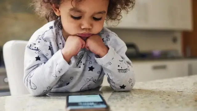 A foto mostra uma criança pequena,bet7k apostascabelos cacheados e pele morena, sentada, com a cabeça apoiada nas mãos e olhando para um celular sobre a mesa. 