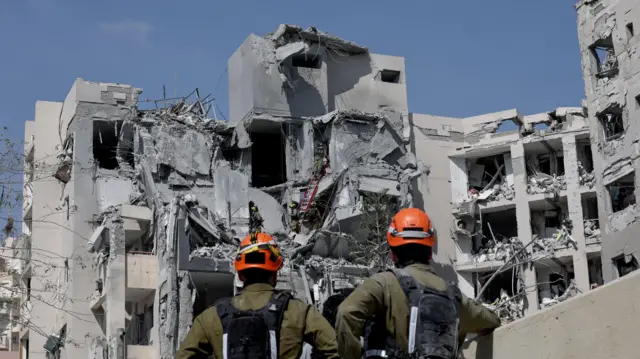 Zdjęcie przedstawia ratowników przed zniszczonym blokiem mieszkalnym.