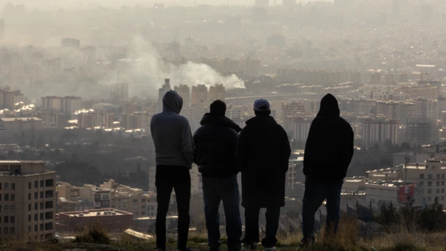 Четверо мужчин вырисовываются на фоне горизонта Тегерана, глядя вниз с холма, как поднимается дым от авиаудара