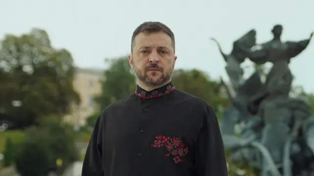 Kadr z przemówienia prezydenta Wołodymyra Zełenskiego. Zełenski, z krótkimi, czarnymi włosami i zarostem, patrzy w kamerę z poważnym wyrazem twarzy. Ma na sobie czarną tunikę z wysokim kołnierzem, ozdobioną czerwono-zielonym wzorem na kołnierzu i lewej stronie klatki piersiowej. 