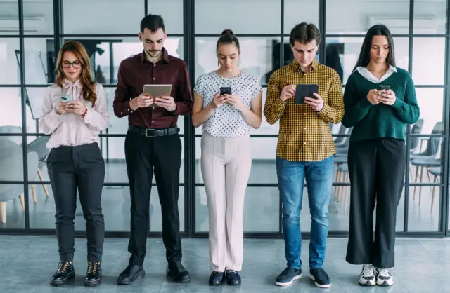 Pięć młodych osób stoi w rzędzie wewnątrz biura, patrząc w ekrany smartfonów i tabletów. Za nimi przeszklona ściana, po drugiej stronie której widoczne są rzędy krzeseł. 