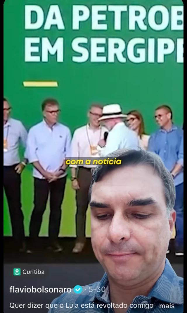 Print de tela de vídeo publicado por Flávio Bolsonaro no X, com ele na frente e um vídeo de Lula ao fundo, em evento público