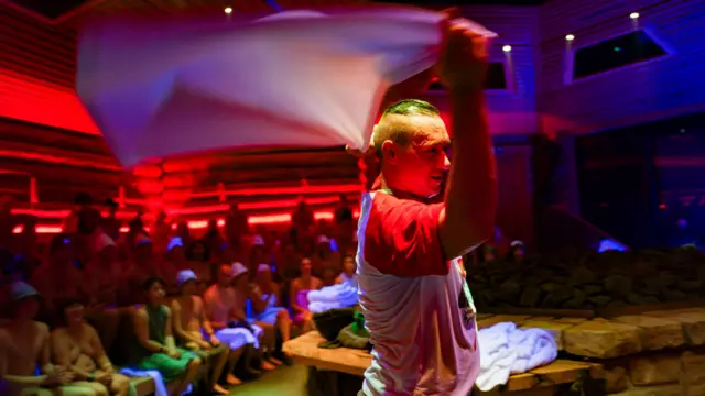 Ludzie siedzą w drewnianej saunie, przyglądając się mężczyźnie, który macha białym ręcznikiem.