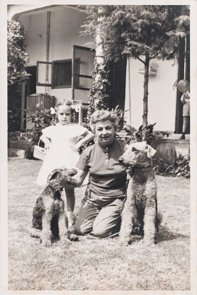 Cristina, con el cabello corto, está vestida con una camisa de manga corta y un pantalón. Está arrodillada sobre la grama de un jardín y ve a la cámara. Con cada mano acaricia a un perro. A su lado está la niña Mara, vestida con un traje blanco. Lleva dos trenzas y en cada una tiene un lazo. Leva una pequeña cartera.