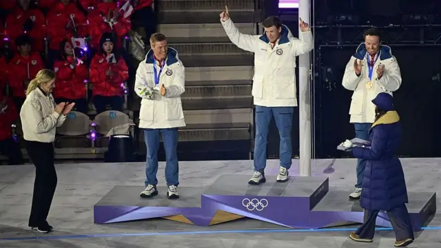 3人が表彰台の上に立っている。真ん中の人は両手を上げ、人差し指を立てている。両脇の2人は手をたたくような仕草で笑顔を見せている。背後に日本選手らが見える